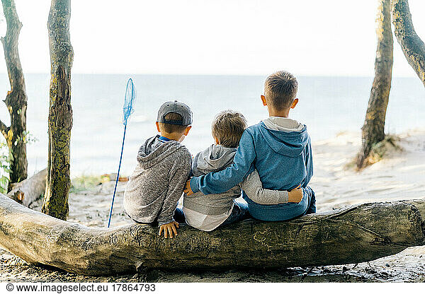 three boys sitting on a log and look at the sea