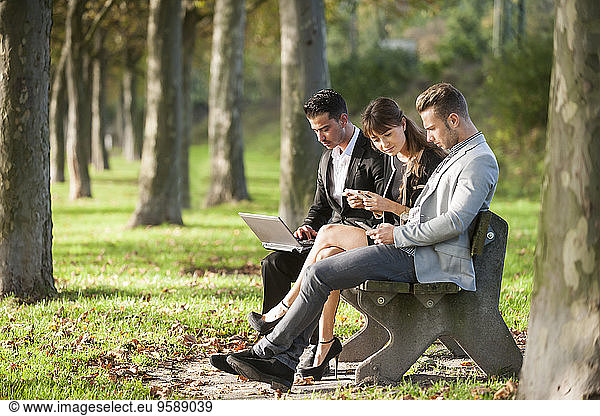 Three business people sitting on a park bench using laptop  smartphone and digital tablet