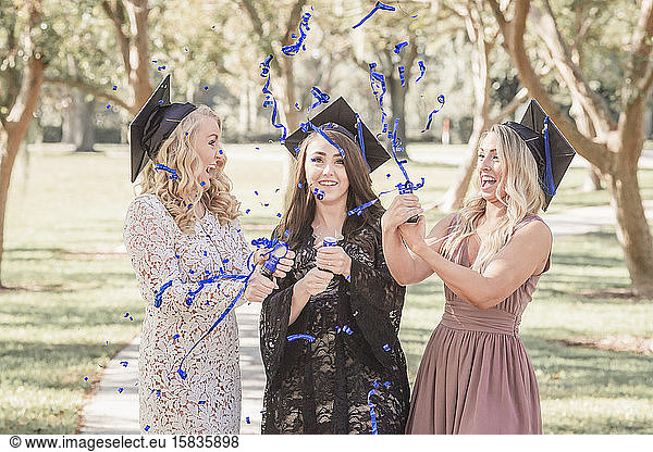 Three caucasian women in graduation caps laughing and popping confetti