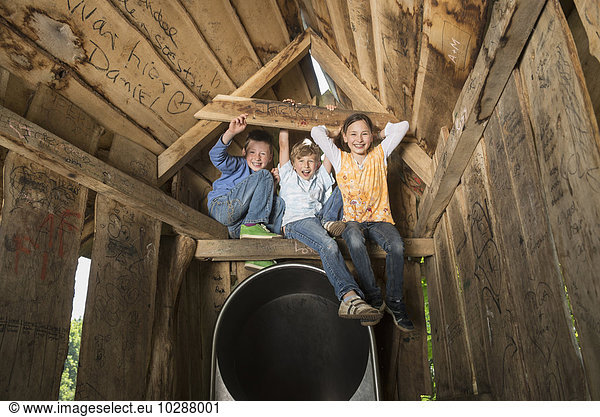 Three children sitting on wooden bar of tree house in a playground  Munich  Bavaria  Germany