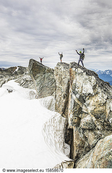 Three climbers stand on a rocky summit with arms raised in the air