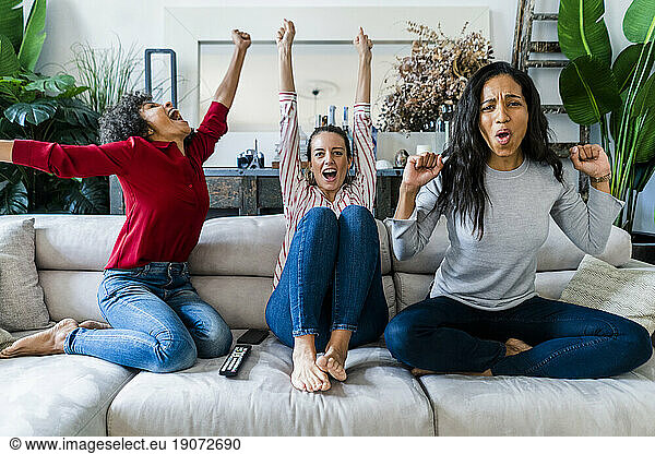 Three excited women on couch at home watching Tv and cheering