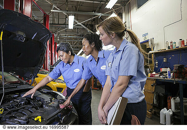 Three female mechanics look inside the engine compartment of a car in a repair shop