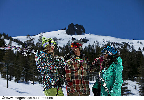 Three friends (one male and two females) hanging out with skis and a snowboard in front of a ski resort.