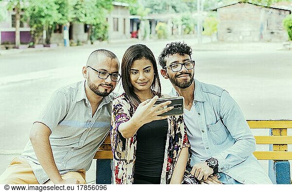 Three friends taking a selfie on a park bench  Three smiling friends sitting on a bench taking a selfie. Front view of three happy friends taking a selfie while sitting on a bench