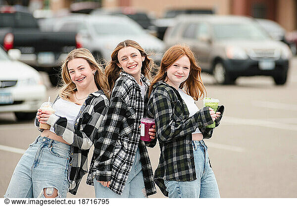 Three happy teen girls holding drinks in a parking lot.