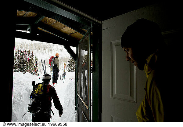 Three male skiers walk out the door of a cabin to go ski touring.