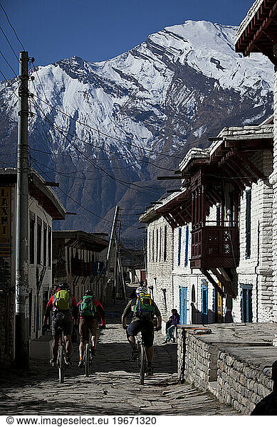 Three men pedal mountain bikes through a small town with tall mountain in background.