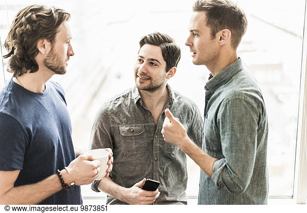 Three men standing talking  one with a cup of coffee  one with a smart phone.