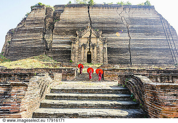 Three monks with orange umbrellas outside a large rock temple  the entrance carved into the rock face at Saigang.