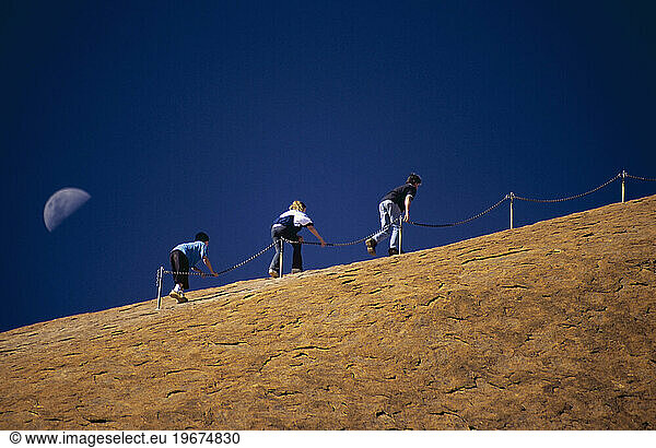 Three people climb Uluru in Australia's Northern Territory  aided by the chains and backed by a half moon.