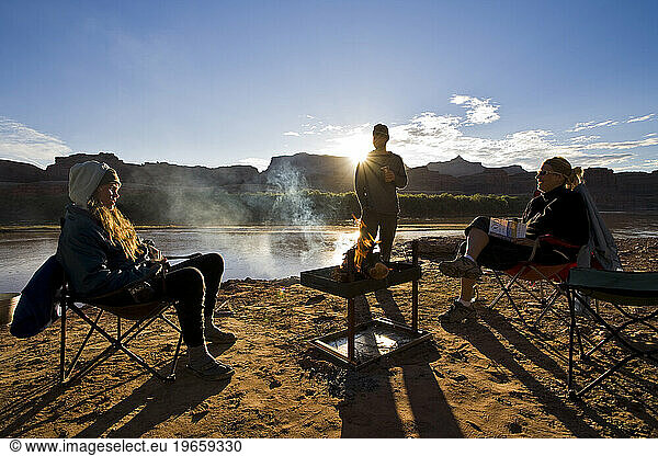 Three people gathered around a fire at sunrise on a sandy beach of the Colorado River in Utah.