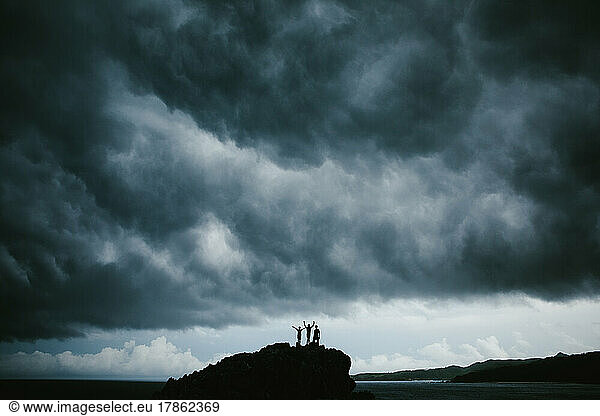 Three people hike on top of a rock with stormy gray sky