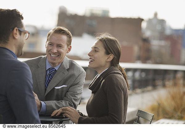 Three people standing in an open space between city buildings  talking to each other. Two men and a woman.