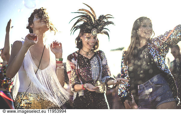 Three smiling young women at a summer music festival face painted  wearing feather headdress  dancing among the crowd.