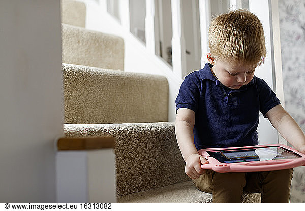 Three year old boy seated on the stairs looking at a digital tablet.