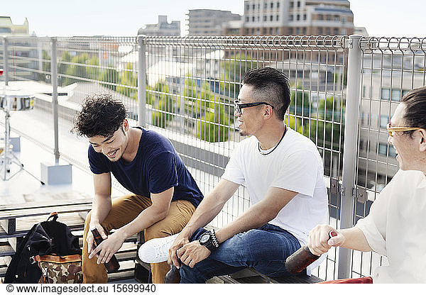 Three young Japanese men sitting on a rooftop in an urban setting  drinking beer.