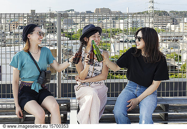 Three young Japanese women sitting on a rooftop in an urban setting  drinking beer.