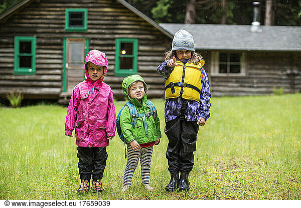 Three young kids in rain gear in front of a log cabin