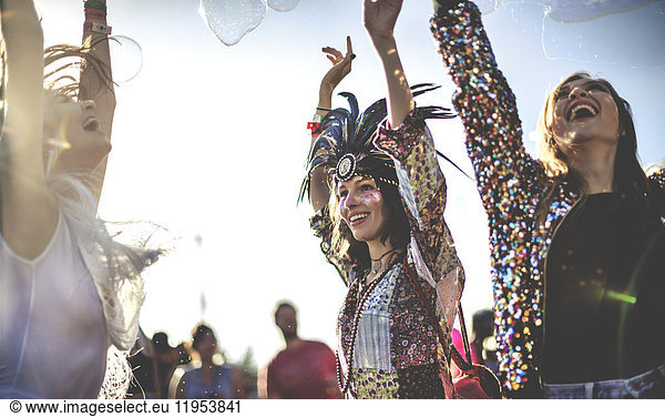 Three young woman at a summer music festival wearing feather headdresses and face paint  arms raised  dancing.