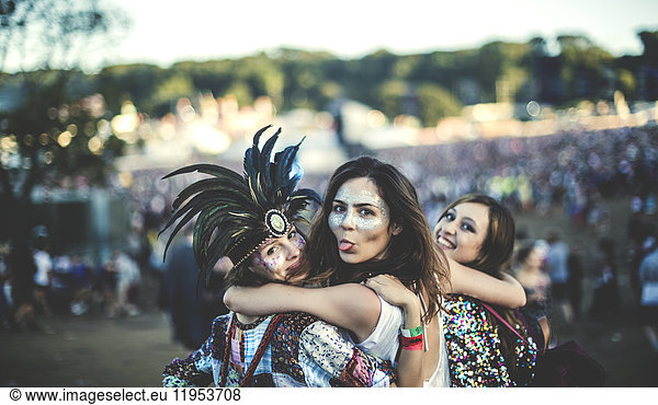 Three young women at a summer music festival feather headdress and faces painted  smiling at camera  sticking out tongue.