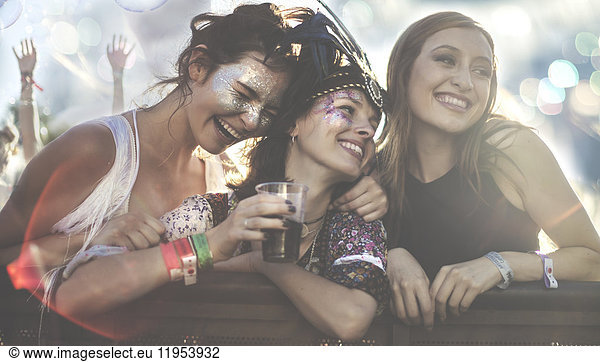 Three young women at a summer music festival wearing feather headdress and faces painted  smiling and holding drink.