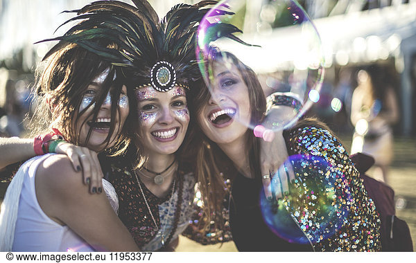 Three young women at a summer music festival wearing feather headdress and faces painted  smiling at camera.