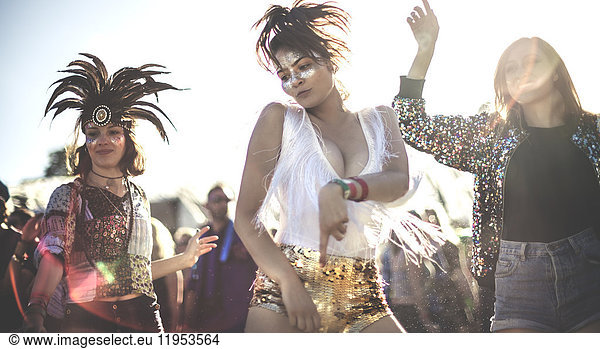 Three young women at a summer music festival wearing sequins and feather headdresses dancing among the crowd.