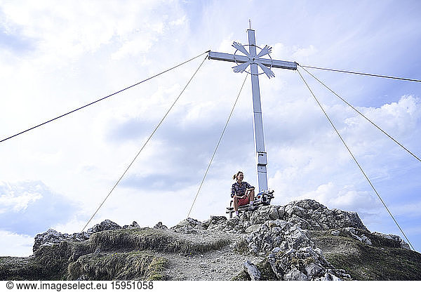 Tiefwinkelansicht einer am Gipfelkreuz des Kramerspitzes sitzenden Frau gegen den Himmel
