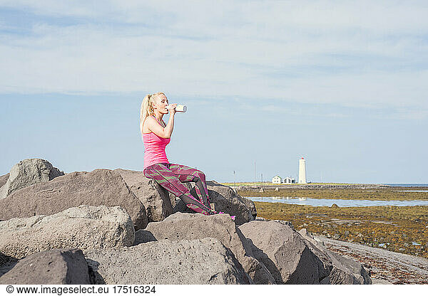 Tired lady drinking water on rocky seashore during workout