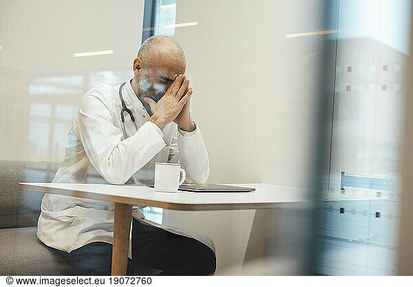 Tired mature doctor sitting with coffee cup and laptop seen through glass
