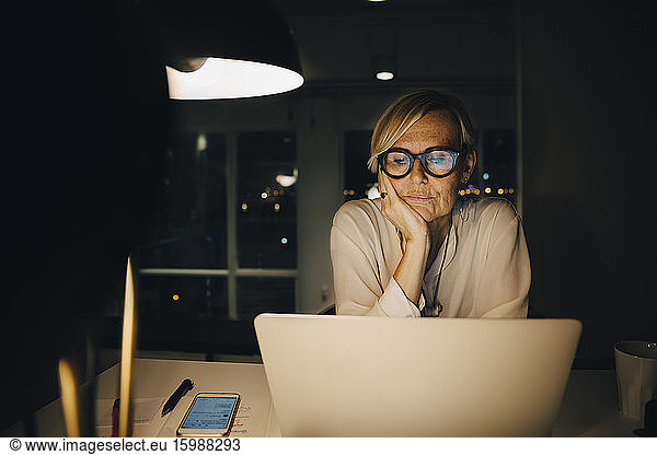 Tired mature female professional staring at laptop while sitting with hand on chin working late in office