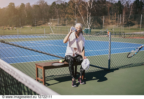 Tired senior woman drinking water while sitting on bench at tennis court