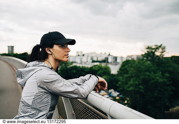 Tired sportswoman looking away while leaning on railing at footbridge in city