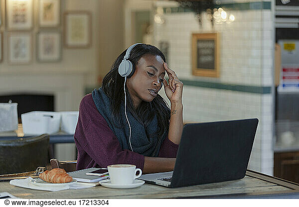 Tired young businesswoman with headphones working at laptop in cafe