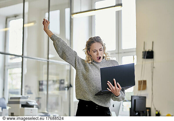 Tired young woman in office holding laptop and yawning