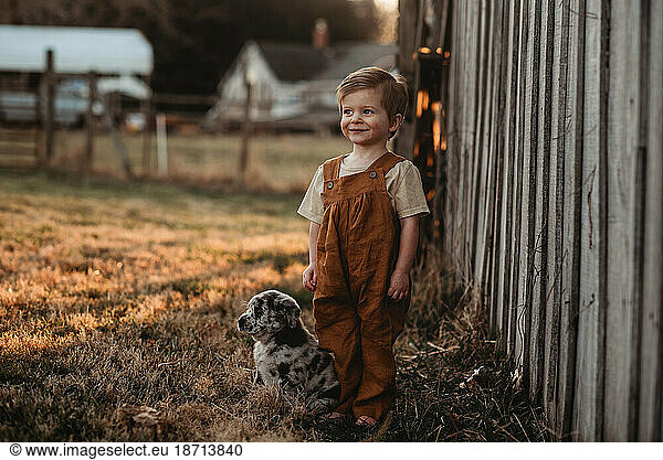 toddler baby boy standing outside by a barn with a puppy