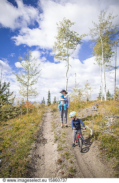 Toddler biking in front of mother carrying baby in forest.