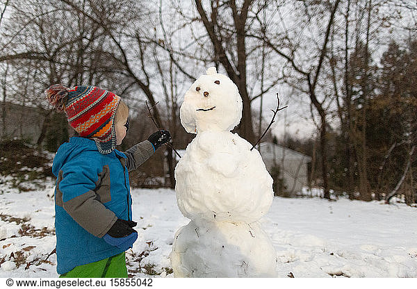 Toddler boy in cold weather gear putting stick arm into a snowman
