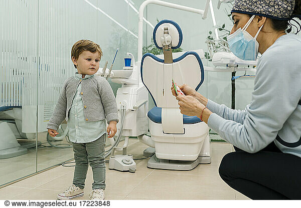 Toddler boy looking at female pediatric dentist with protective face mask in clinic
