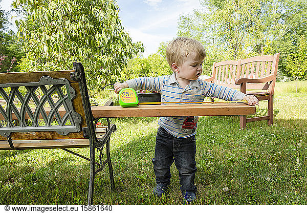 Toddler boy looks serious as he measures a board outside in backyard