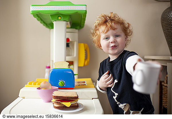 Toddler boy offering a cup of coffee in front of his play kitchen