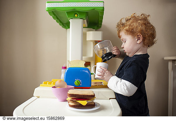Toddler boy playing in toy kitchen pretending to pour coffee into cup