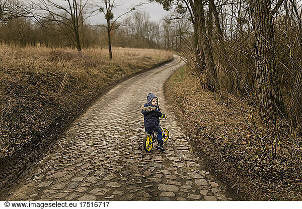 Toddler boy riding push bike on country road in warm hat