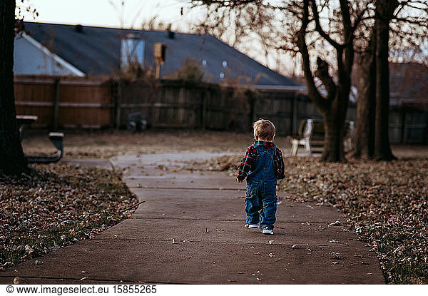 Toddler boy wearing overalls walks through park on fall day.