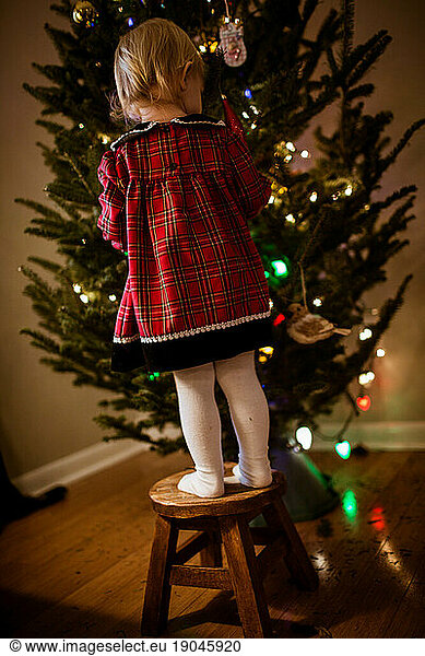 toddler girl standing wearing plaid dress in front of christmas tree