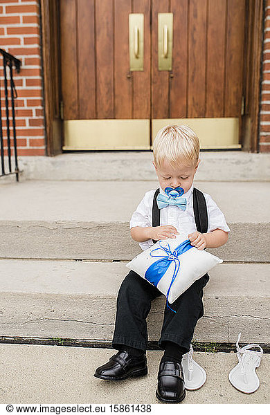 Toddler ring bearer with pacifier sits on steps of church