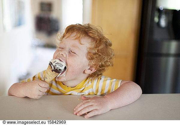 Toddler taking a big bite of his ice cream come at home in kitchen
