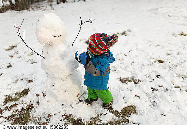 Toddler wearing cold weather gear stands next to snowman holding snow