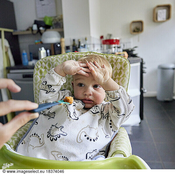 Toddler with hands up refusing food sitting in high chair in kitchen